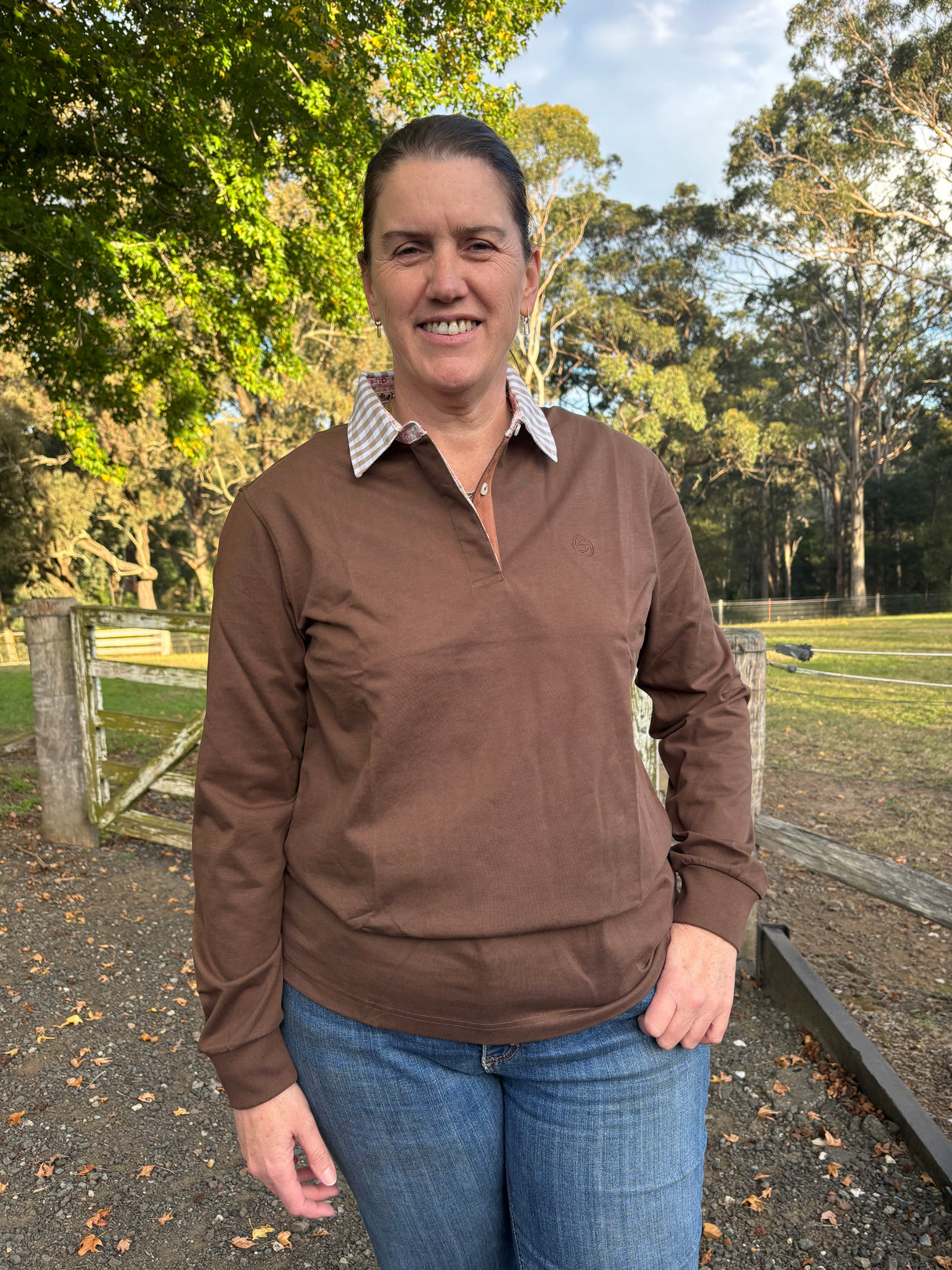 Woman wearing a chocolate brown long-sleeve rugby shirt and blue jeans standing outdoors with trees in the background
