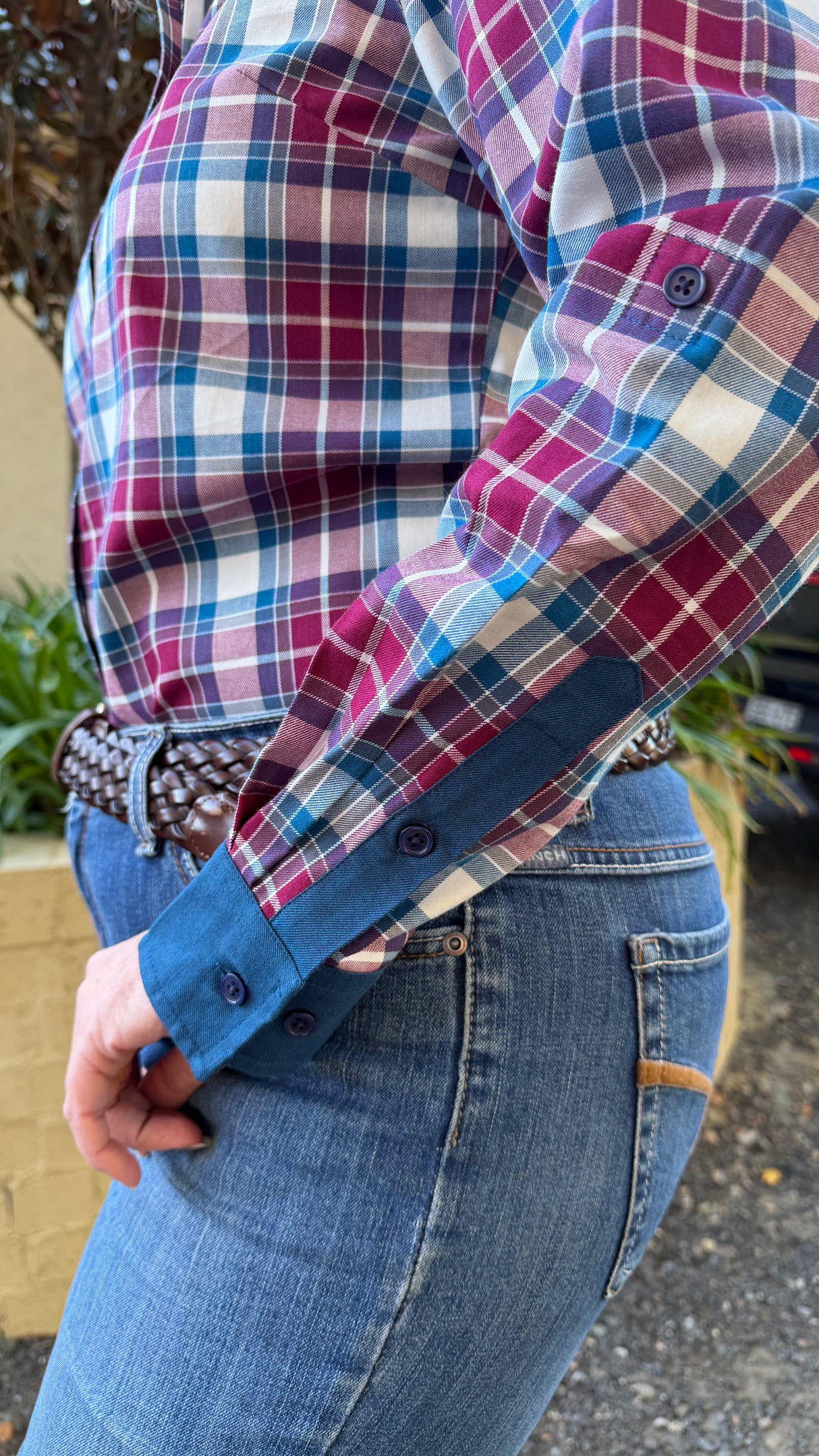 A close-up of a woman wearing a long-sleeve plaid button-up shirt with blue contrast patch pockets and blue jeans, standing outdoors.