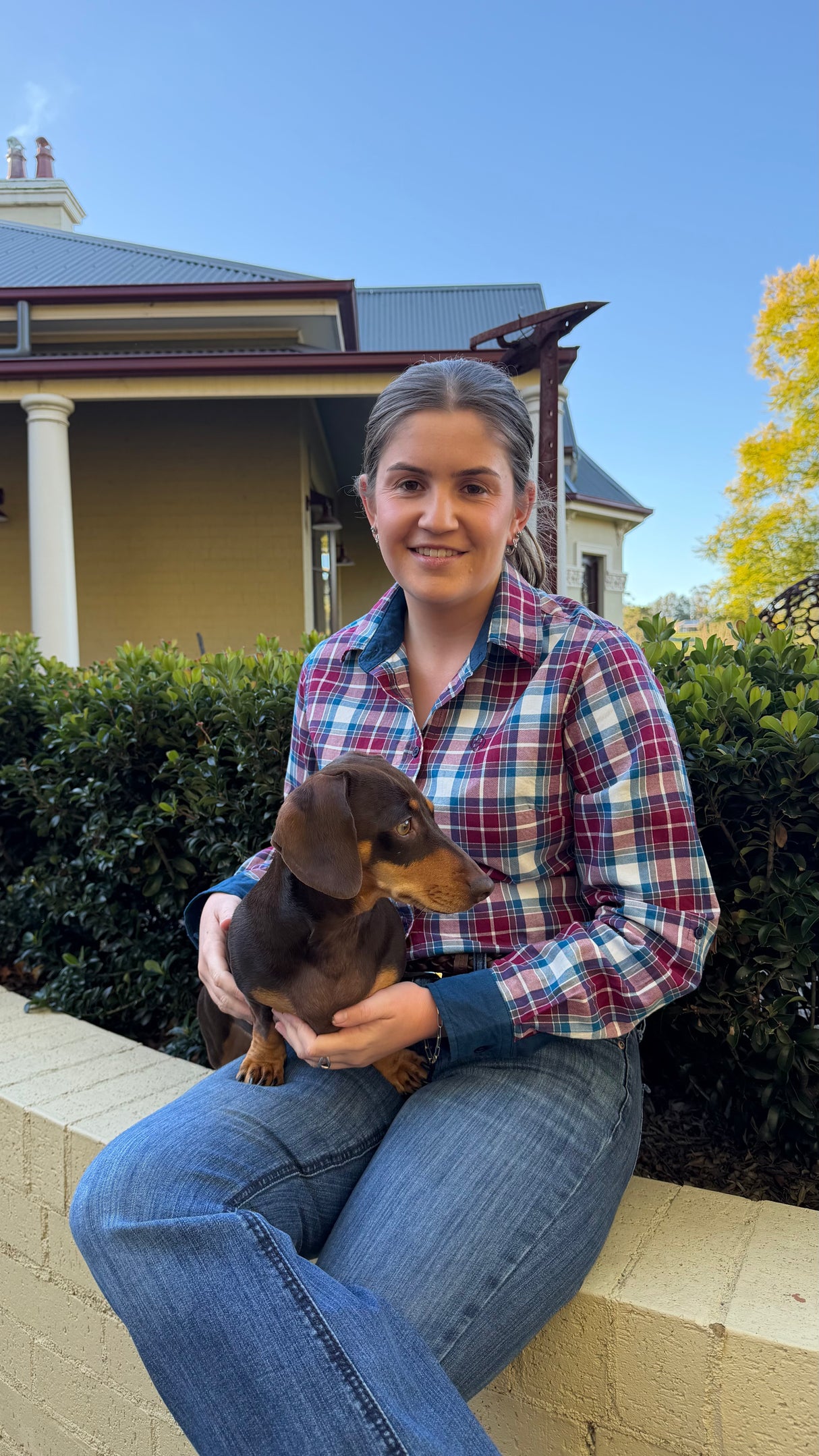 A woman sitting outdoors on a low wall holding a small brown dachshund puppy. She is wearing a red, blue, and white plaid button-up shirt and blue jeans, with a house, hedges, and blue sky in the background.