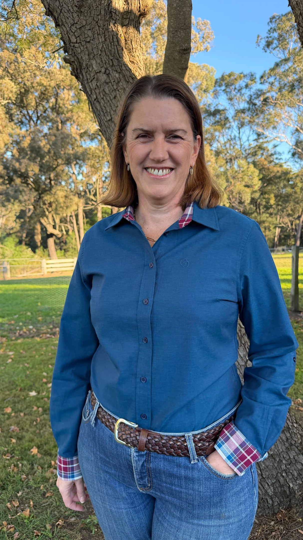 A smiling woman standing outdoors in front of a tree, wearing a blue long-sleeve button-up shirt with a plaid collar and cuff, blue jeans, and a brown braided belt.
