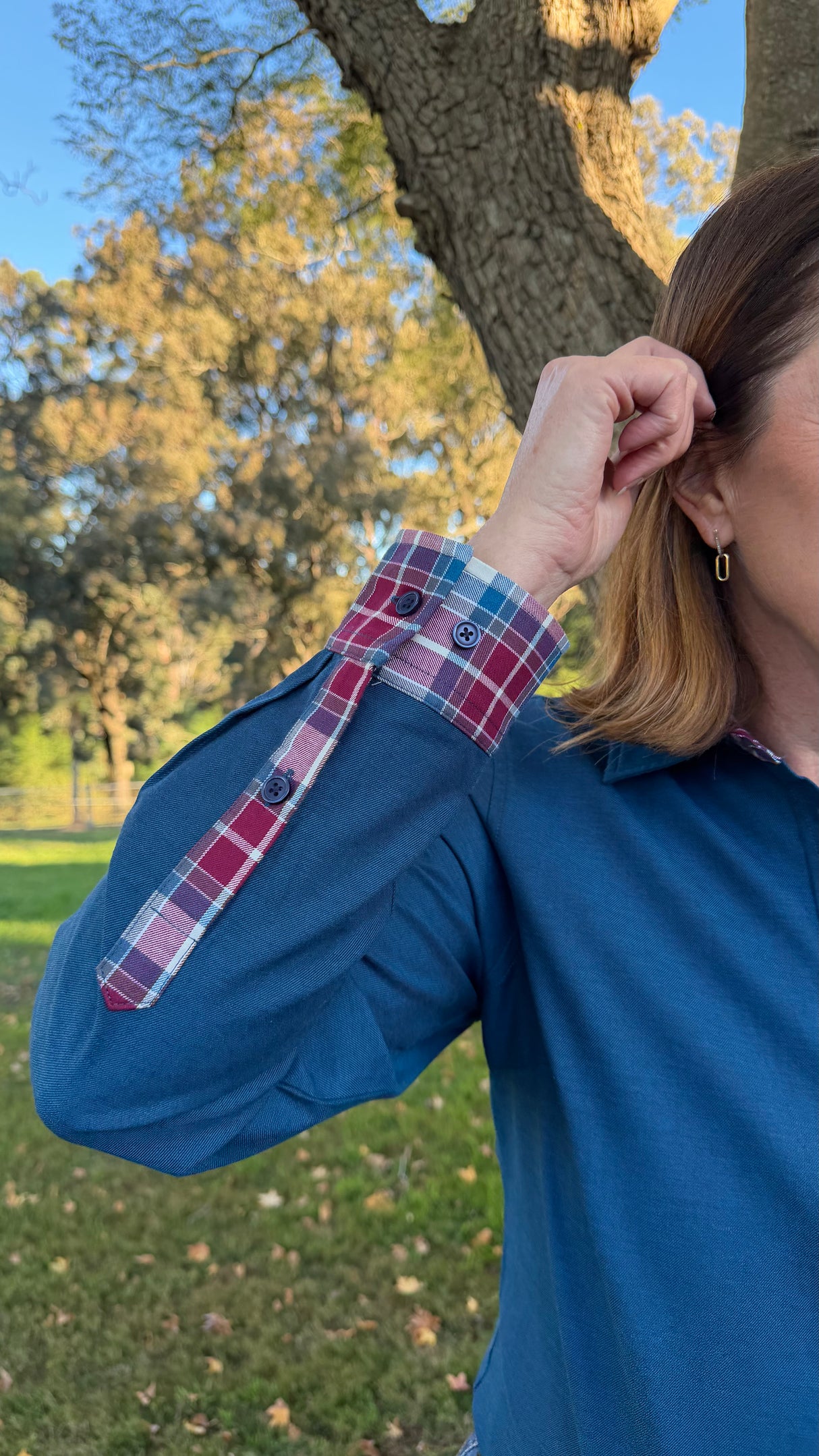 A woman outdoors wearing a blue long-sleeve top with a red, blue, and white plaid button-up cuff, with a tree and grassy park in the background.