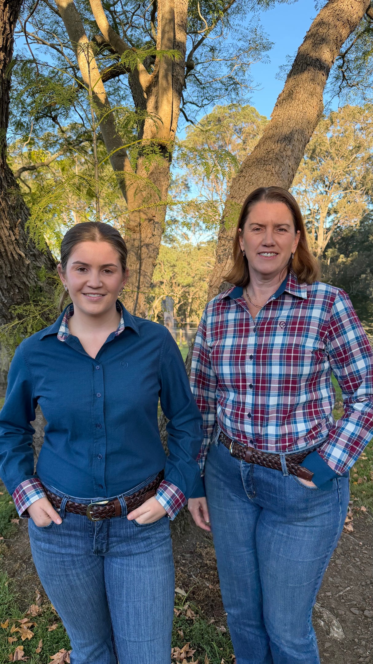 Two women standing outdoors in front of large trees, wearing long-sleeve button-up shirts and blue jeans. The woman on the left wears a solid blue shirt, and the woman on the right wears a red, white, and blue plaid shirt.