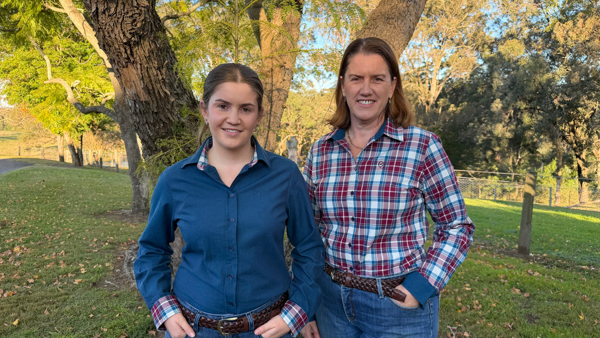 Two women standing outdoors in front of trees, wearing long-sleeve button-up shirts. The woman on the left wears a solid dark blue shirt, and the woman on the right wears a red, white, and blue plaid shirt.