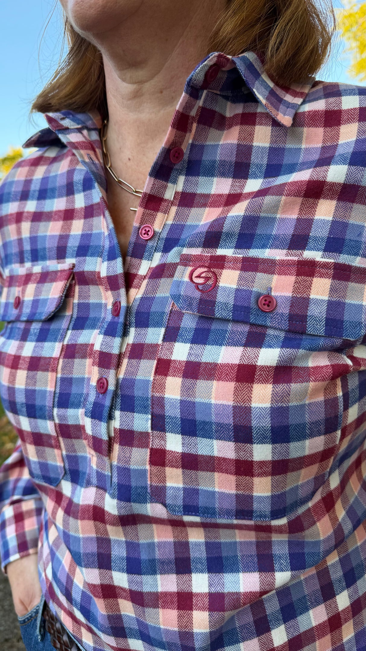 Close-up of a person wearing a plaid button-up shirt in blue, purple, pink, and white, with a small embroidered logo on the chest pocket.