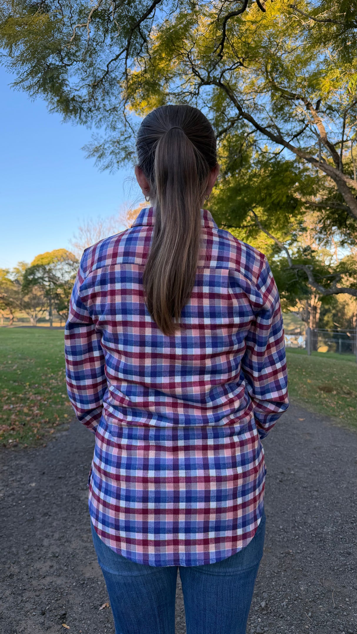 A woman viewed from behind wearing a red, blue, and white plaid button-up shirt and blue jeans, standing on a dirt path in a park with trees and blue sky in the background.