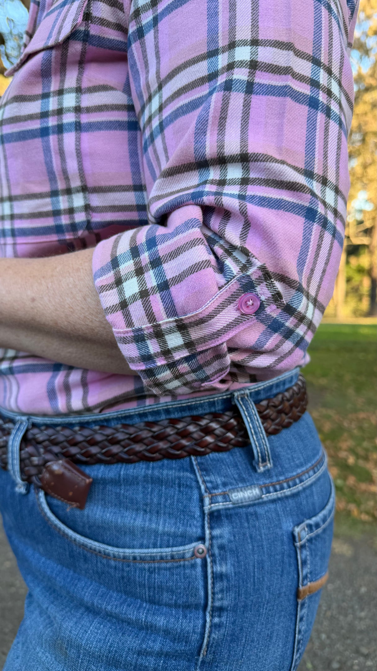 A close-up lifestyle shot of a person wearing a pink and blue plaid button-down shirt, rolled-up sleeves, and a brown braided belt with blue jeans, outdoors.