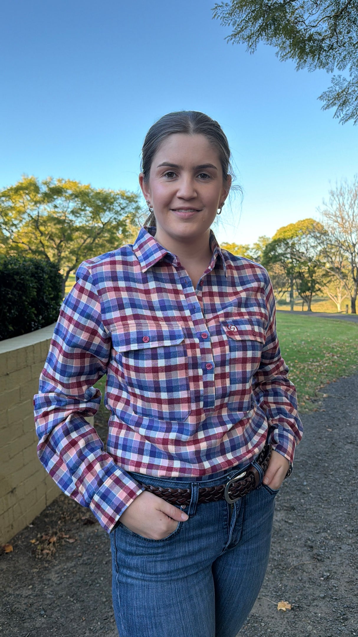 A woman wearing a red, blue, and white plaid button-down shirt and blue jeans, standing outdoors on a path with trees and grass in the background.