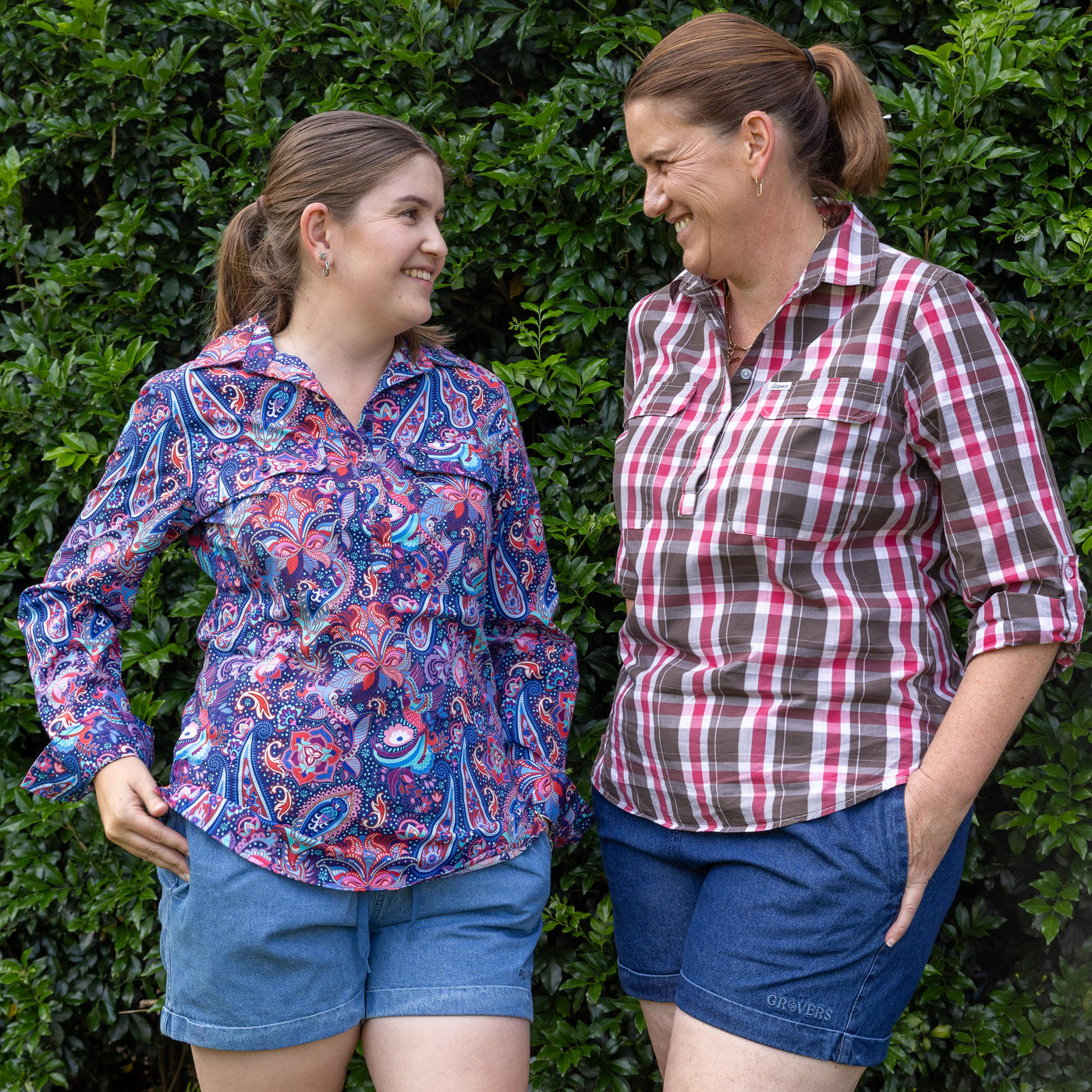 Two women standing outdoors against a green hedge, wearing patterned shirts and denim shorts.