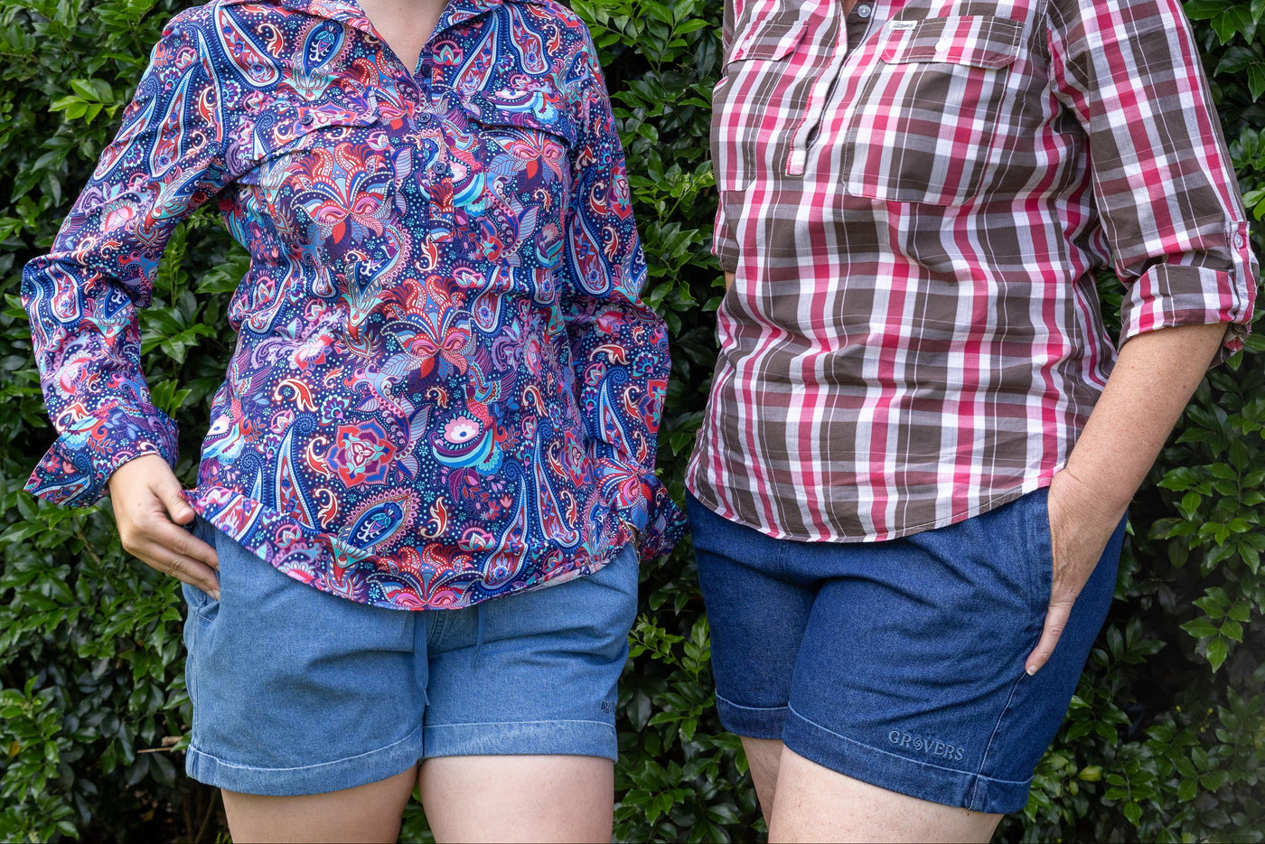 Two women standing outdoors against a green hedge, wearing cotton shirts patterned with floral, paisley and check with denim shorts.