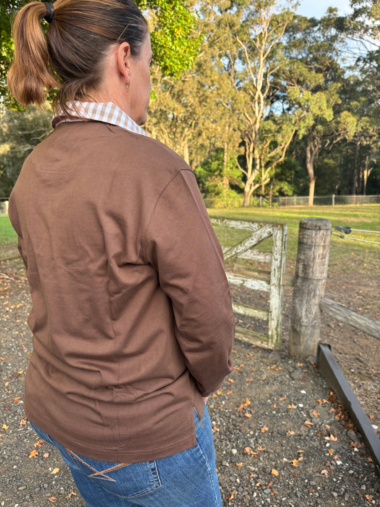 Woman wearing a chocolate brown rugby and blue jeans standing in an outdoor setting with trees and a wooden gate.