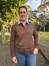 Woman wearing a chocolate brown long-sleeve rugby shirt and blue jeans standing outdoors with trees in the background