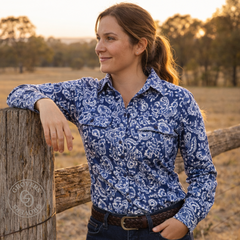 Woman wearing a blue patterned shirt standing by a wooden fence in a field.