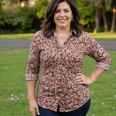 Woman wearing a patterned shirt untucked over  jeans standing outdoors with greenery in the background