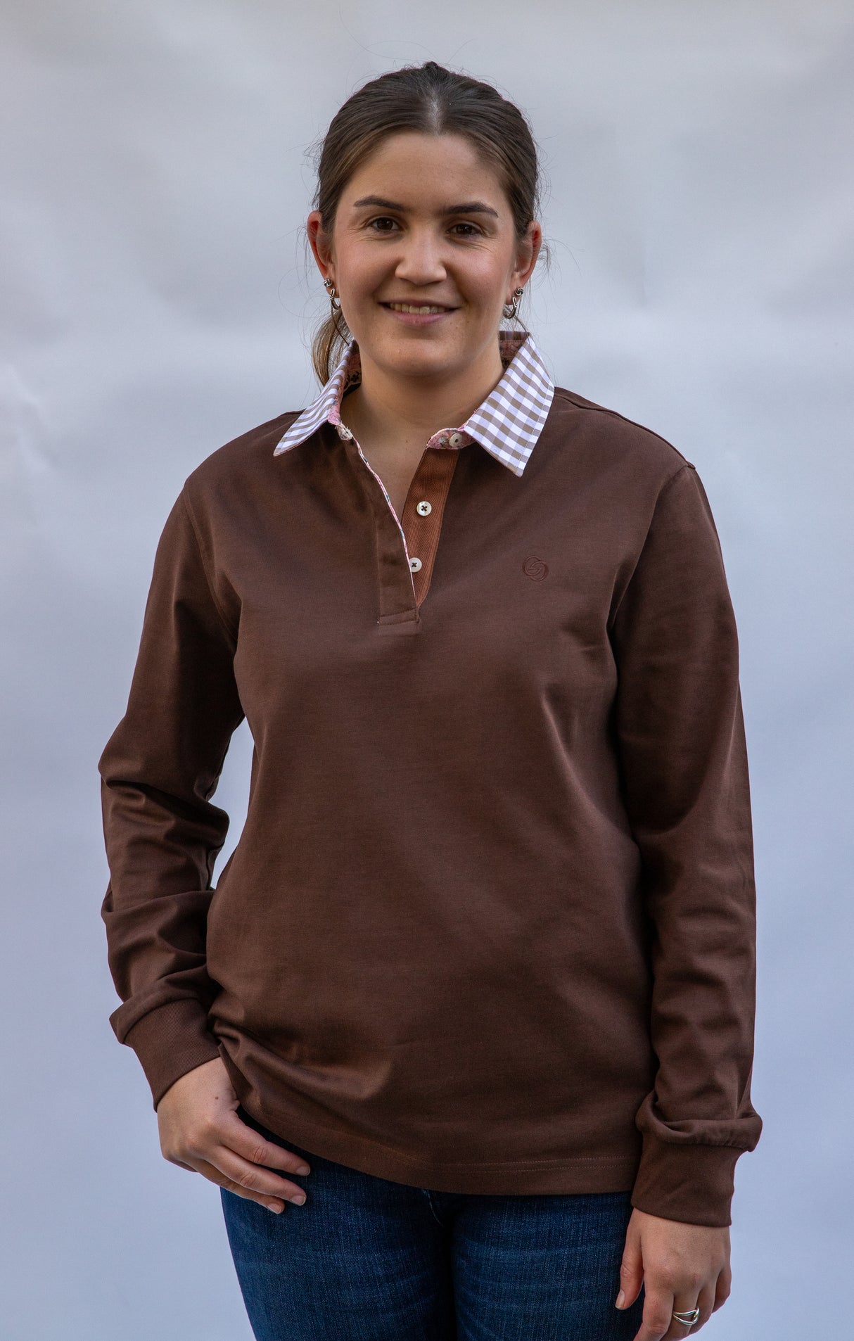 Young Woman wearing a chocolate brown long-sleeve rugby shirt with a gingham collar against a plain background