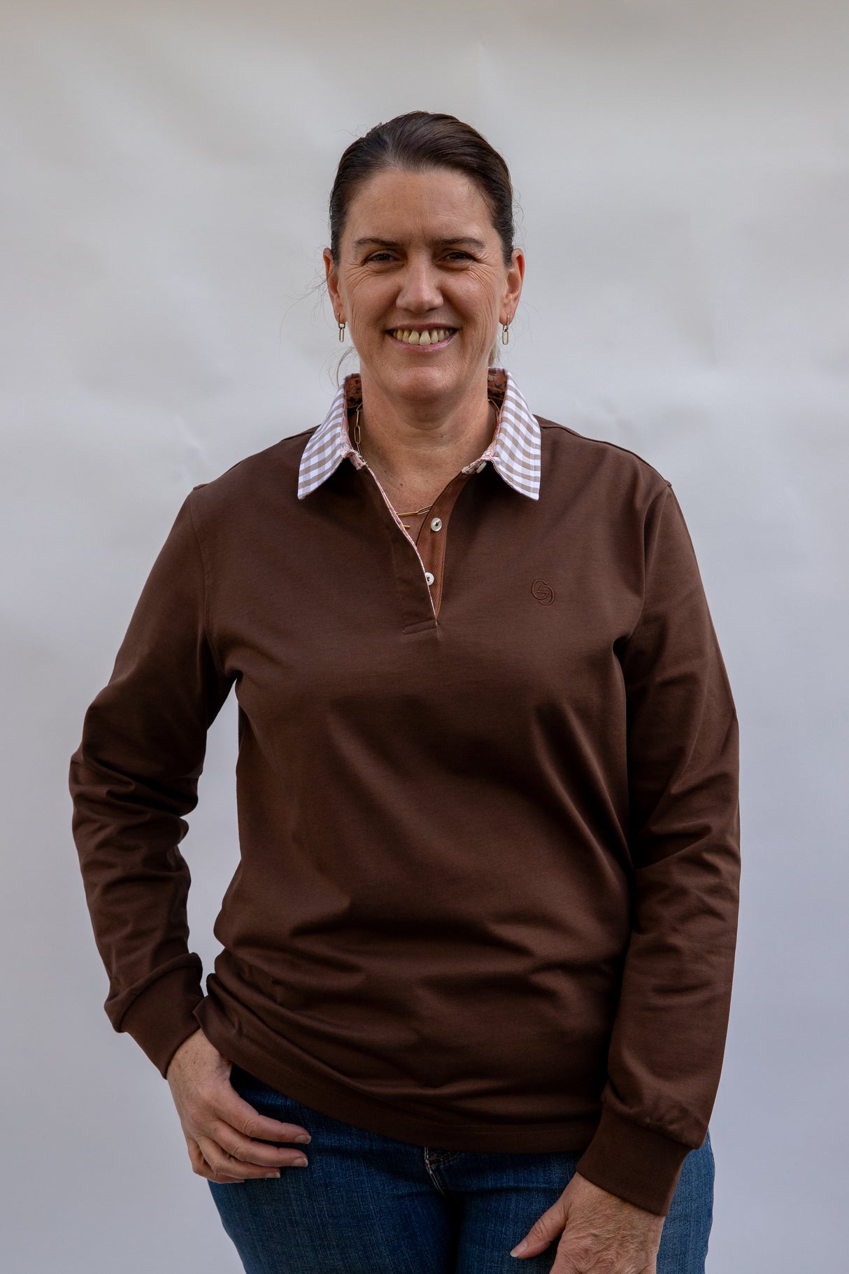 middle aged Woman wearing a chocolate brown long-sleeve rugby shirt with a gingham collar against a plain background