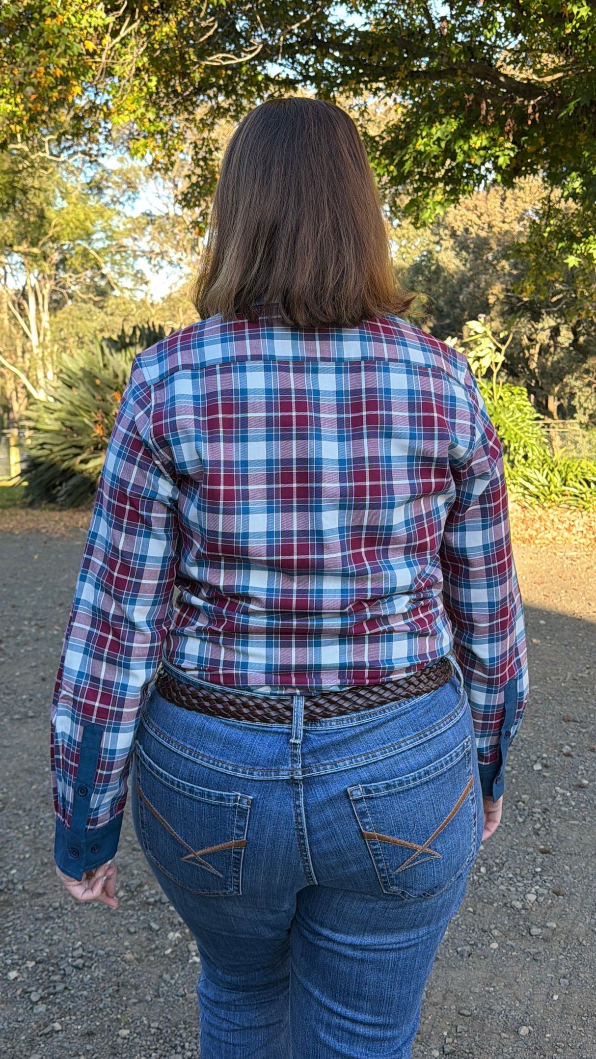 A woman viewed from behind wearing a red, blue, and white plaid long-sleeve button-up shirt and blue jeans, standing outdoors on a gravel path with trees in the background.