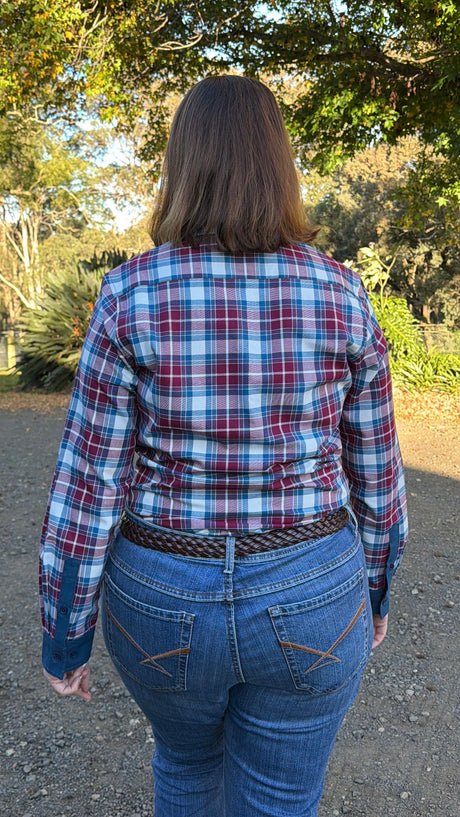 A woman viewed from behind wearing a red, blue, and white plaid long-sleeve button-up shirt and blue jeans, standing outdoors on a gravel path with trees in the background.