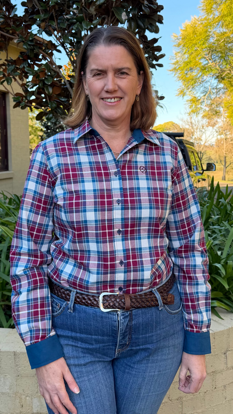 A woman wearing a red, blue, and white plaid long-sleeve button-up shirt with blue jeans, standing outdoors in front of a house and trees.