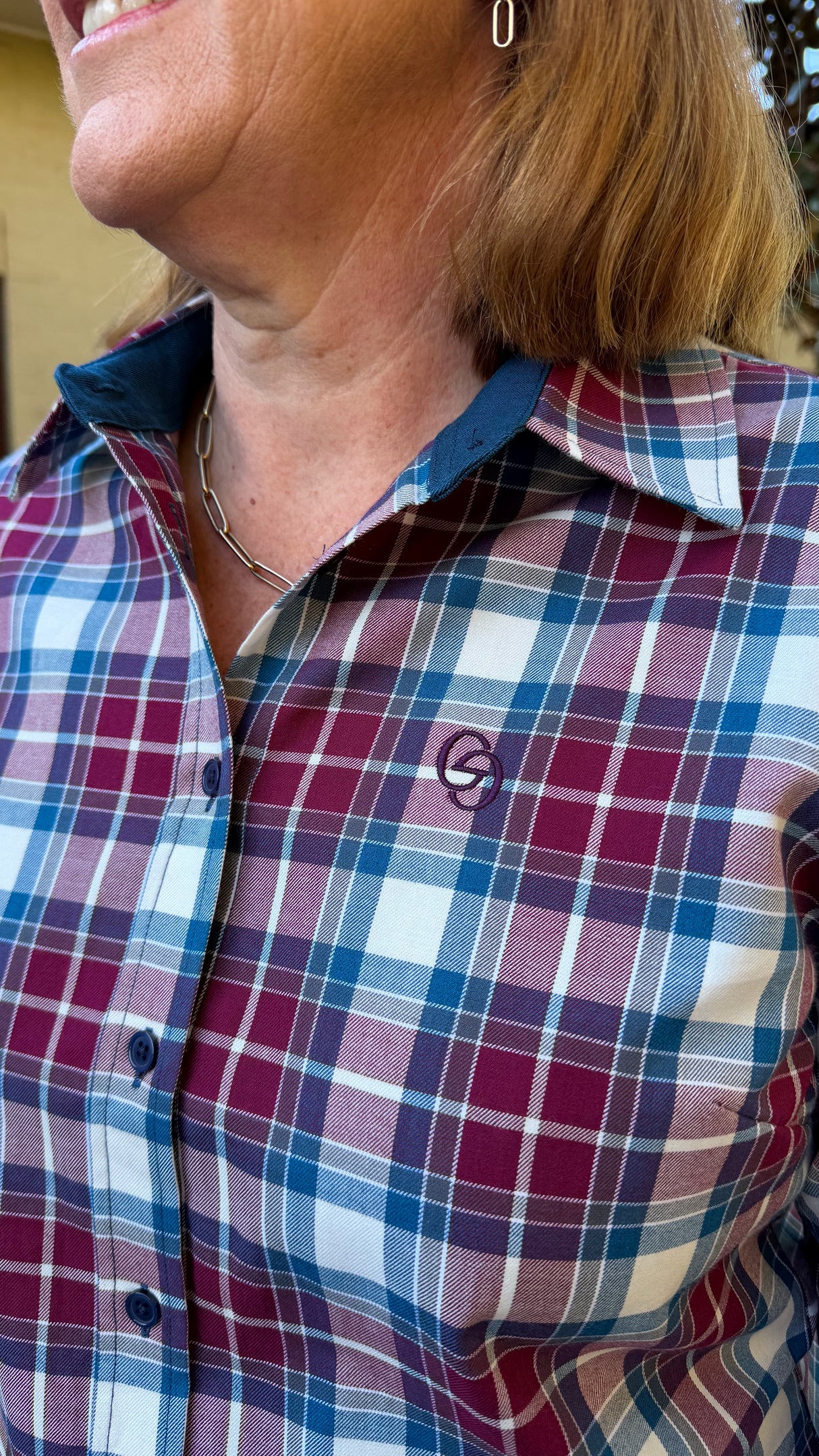 Close-up of a woman wearing a plaid button-up shirt in burgundy, blue, and white, with a small embroidered circular logo on the chest.