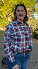 A woman smiling outdoors wearing a red, blue, and white plaid long-sleeve button-up shirt with blue jeans and a brown belt, standing on a dirt path with trees in the background.