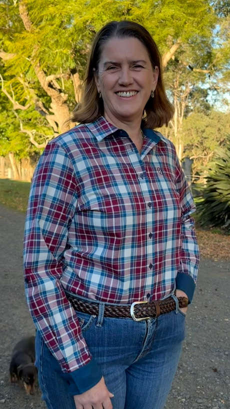 A woman smiling outdoors wearing a red, blue, and white plaid long-sleeve button-up shirt with blue jeans and a brown belt, standing on a dirt path with trees in the background.