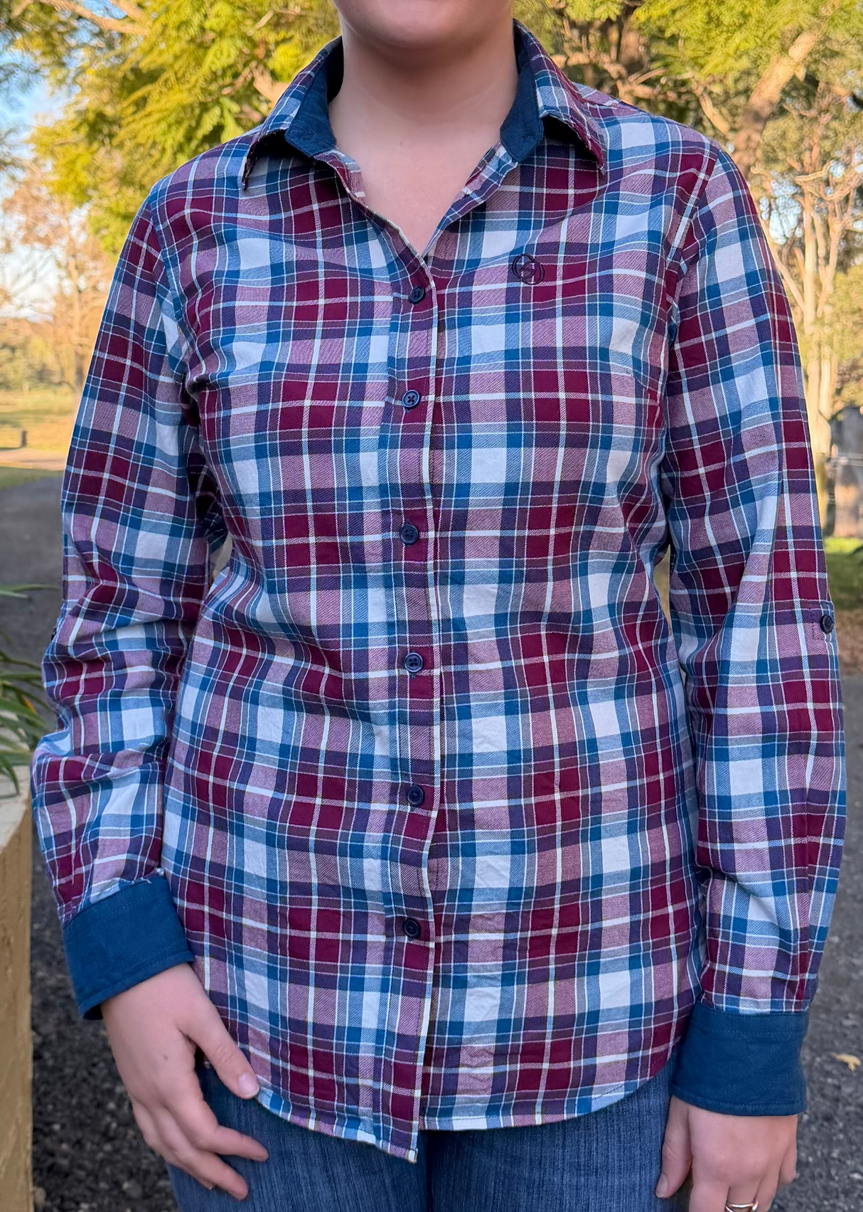 A person wearing a long-sleeve plaid button-up shirt in red, blue, and white, paired with blue jeans, photographed outdoors in a park setting.