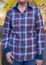 A person wearing a long-sleeve plaid button-up shirt in red, blue, and white, paired with blue jeans, photographed outdoors in a park setting.