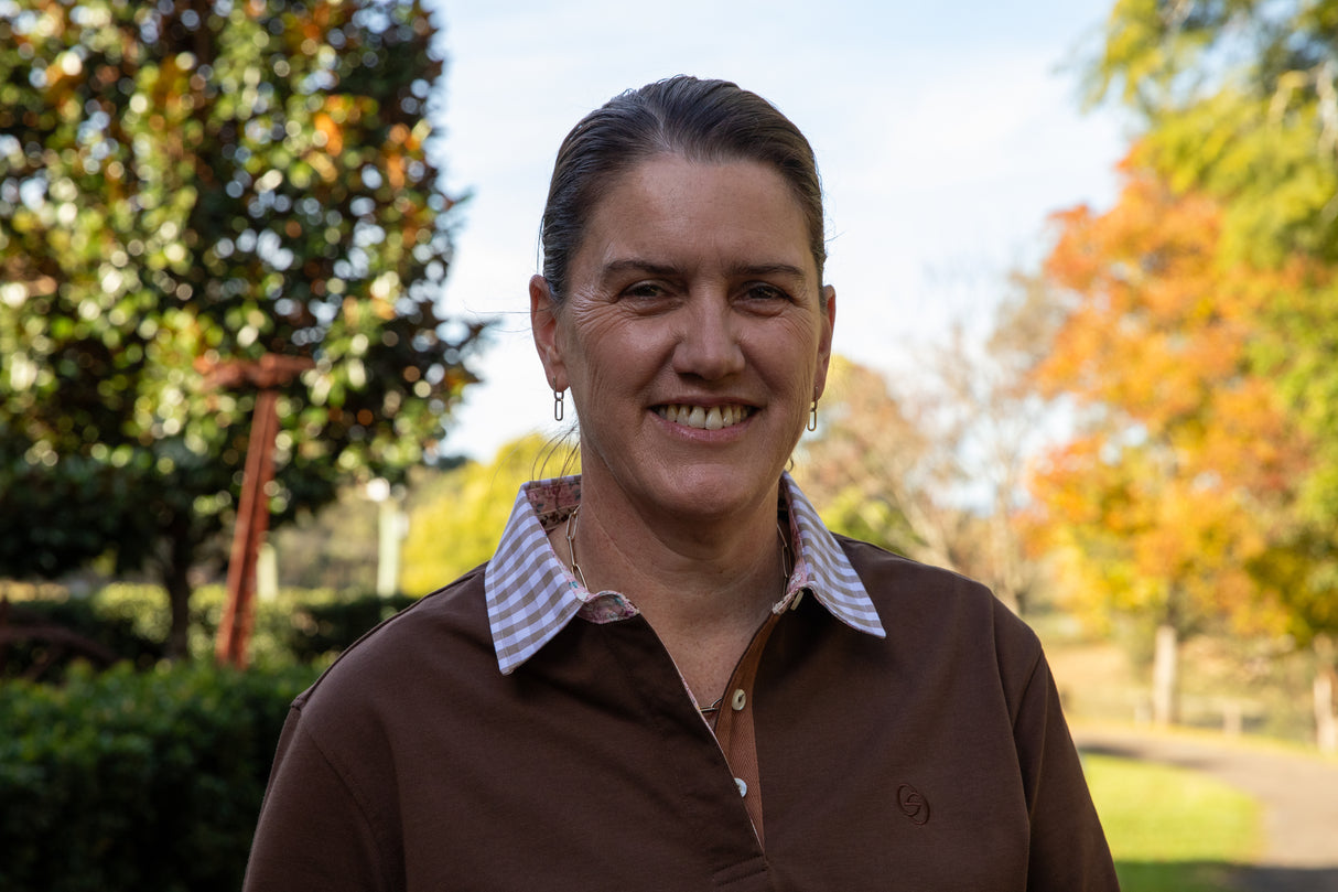 Woman wearing a chocolate brown rugby shirt standing outdoors with trees and a clear sky in the background