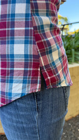 Close-up of a person wearing a red, blue, and white plaid shirt , showing side splits over  blue jeans, with an outdoor background visible.