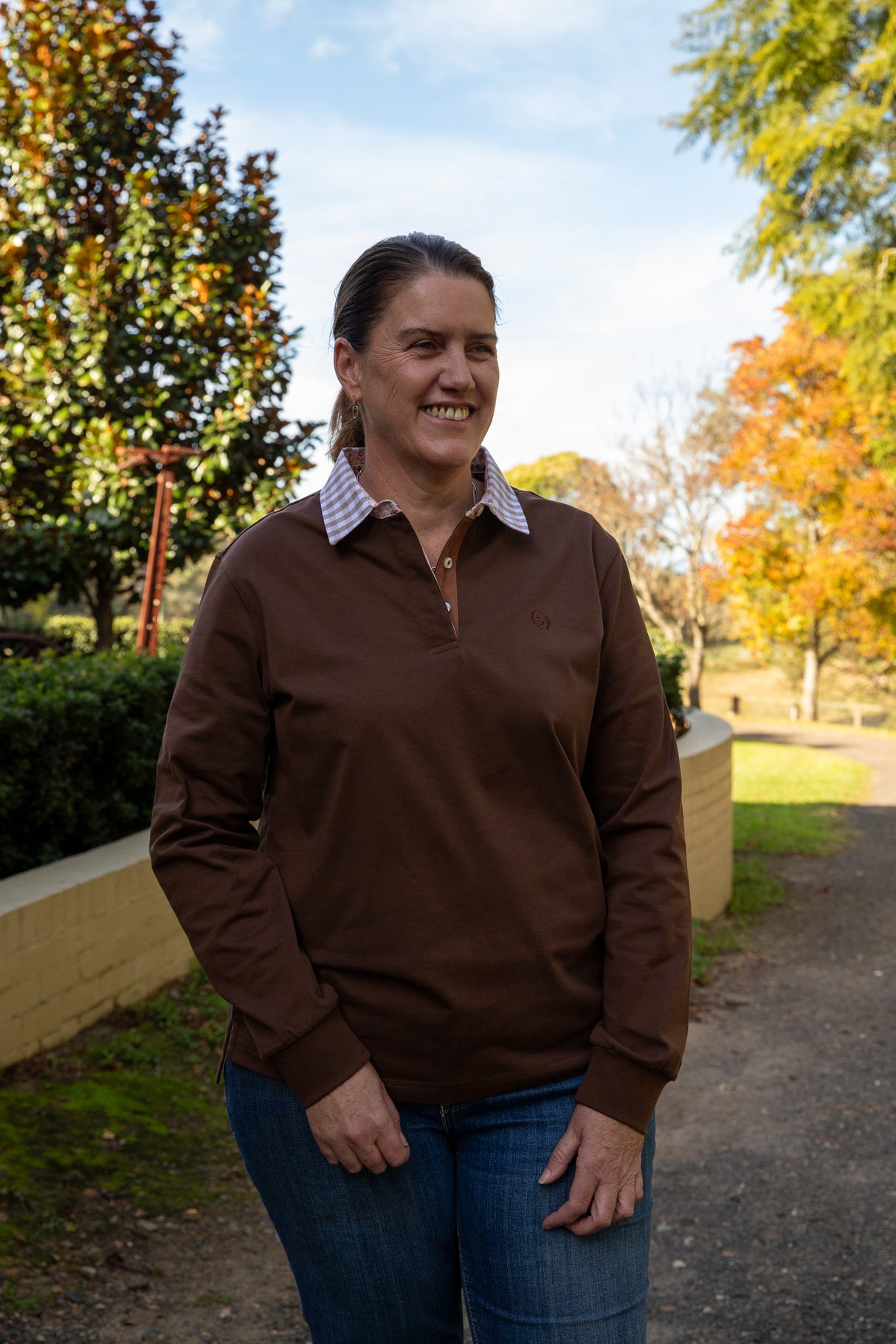 middle aged woman wearing a chocolate brown rugby with a gingham collar, standing outdoors with trees and a clear sky in the background 