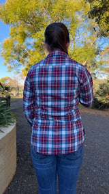 A person viewed from behind wearing a red, white, and blue plaid button-up shirt and blue jeans, standing outdoors on a paved path with trees and greenery in the background.