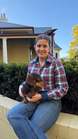 A woman sitting outdoors on a low wall holding a small brown dachshund puppy. She is wearing a red, blue, and white plaid button-up shirt and blue jeans, with a house, hedges, and blue sky in the background.