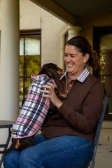 Woman wearing chocolate brown rugby shirt and blue jeans sitting on a verandah holding a small dog wearing a plaid coat.