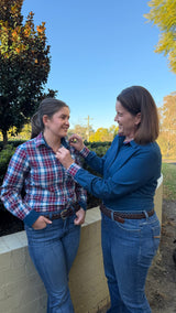 Two women standing outdoors near a low wall, one adjusting the collar of the other woman's plaid button-up shirt. The woman on the left wears a red, white, and blue plaid shirt with blue jeans, and the woman on the right wears a blue long-sleeve top and blue jeans.