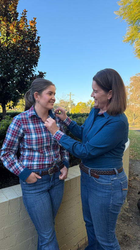 Two women standing outdoors near a low wall, one adjusting the collar of the other woman's plaid button-up shirt. The woman on the left wears a red, white, and blue plaid shirt with blue jeans, and the woman on the right wears a blue long-sleeve top and blue jeans.