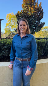 A woman wearing a blue long-sleeve button-up shirt and blue jeans, standing outdoors in front of a beige brick wall, green hedges, and trees under a clear blue sky.