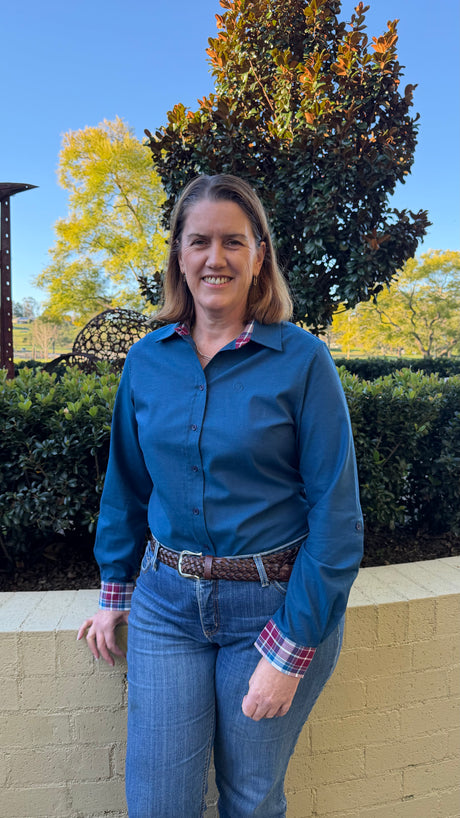 A woman wearing a blue long-sleeve button-up shirt and blue jeans, standing outdoors in front of a beige brick wall, green hedges, and trees under a clear blue sky.