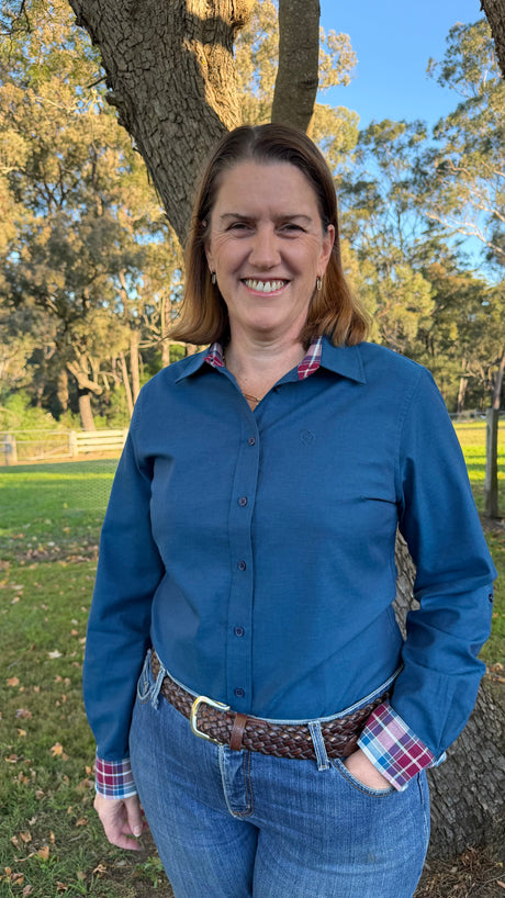 A smiling woman standing outdoors in front of a tree, wearing a blue long-sleeve button-up shirt with a plaid collar and cuff, blue jeans, and a brown braided belt.