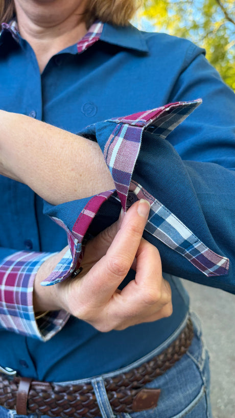 A close-up of a person wearing a blue button-up shirt with a plaid lining, showing the inner plaid cuff as they roll up the sleeve. The person is also wearing a brown braided belt and blue jeans.