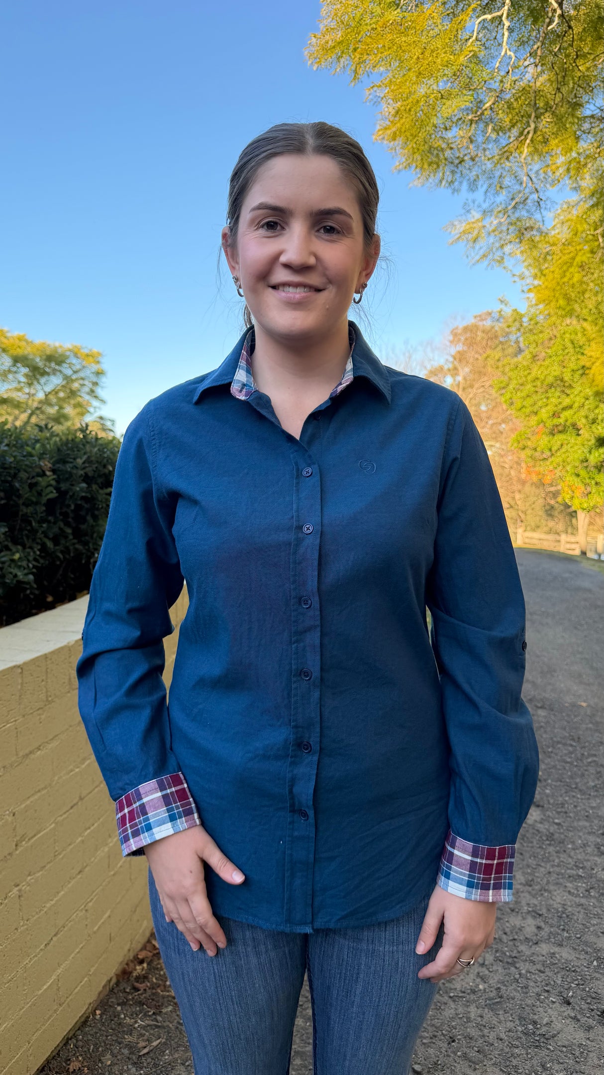 A woman wearing a dark blue long-sleeve button-up shirt with a red, blue, and white plaid cuff lining, standing outdoors on a paved path with trees and a yellow wall in the background.