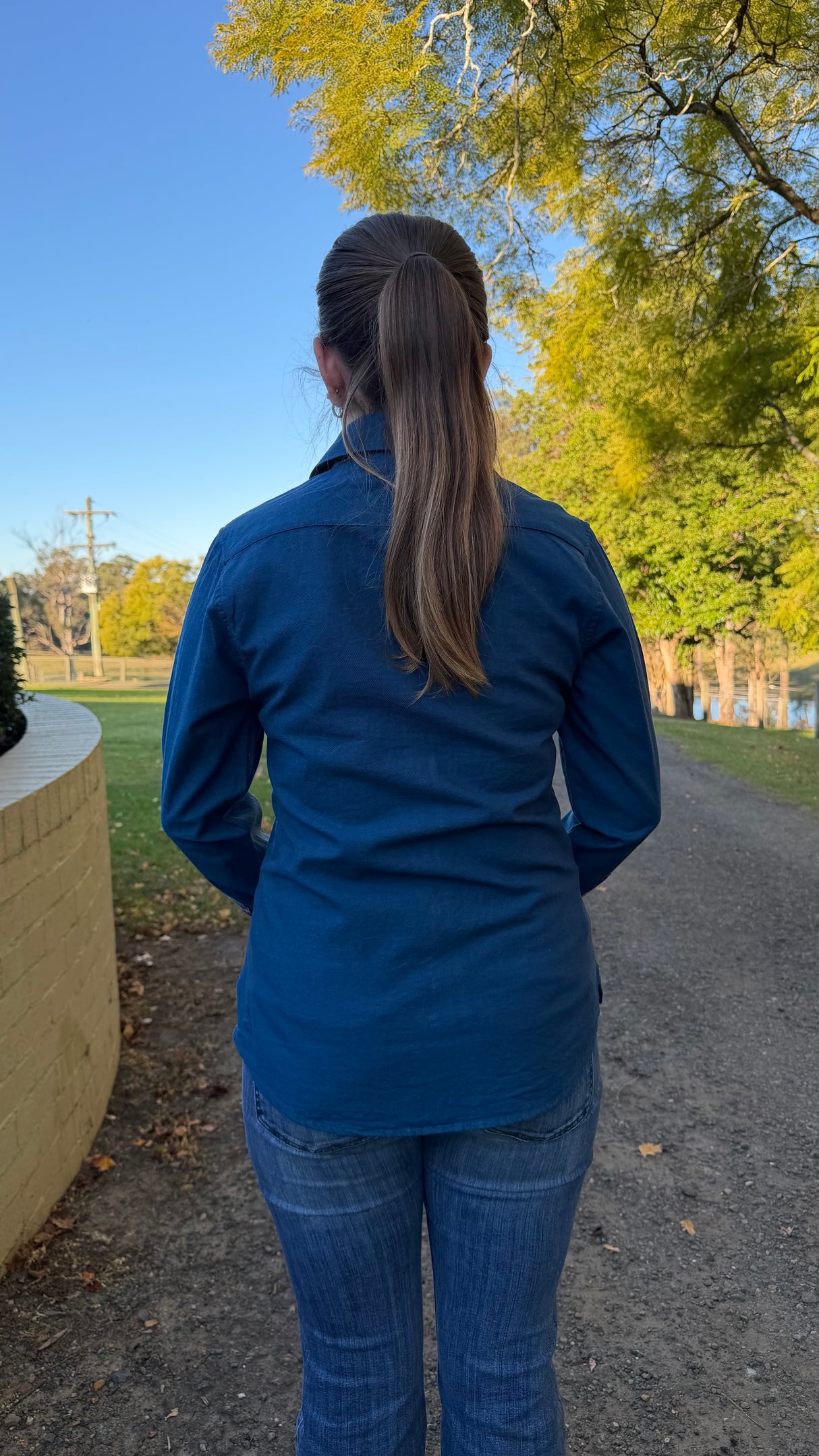 A woman viewed from behind wearing a dark blue long-sleeve button-up shirt and blue jeans, standing on a gravel path outdoors with trees and blue sky in the background.