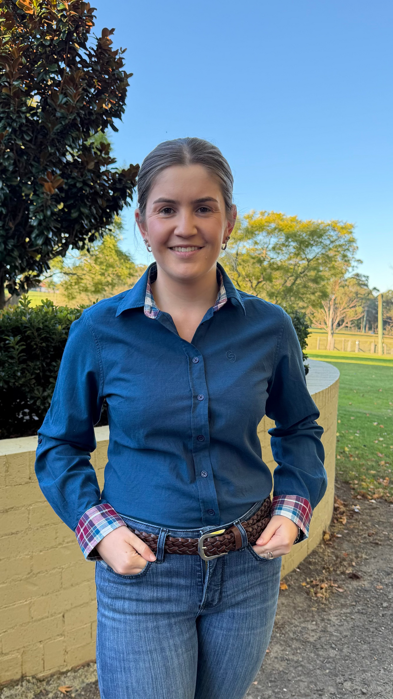 A woman wearing a blue long-sleeve button-up shirt and blue jeans, standing outdoors in front of a yellow brick wall, green grass, and trees under a clear blue sky.
