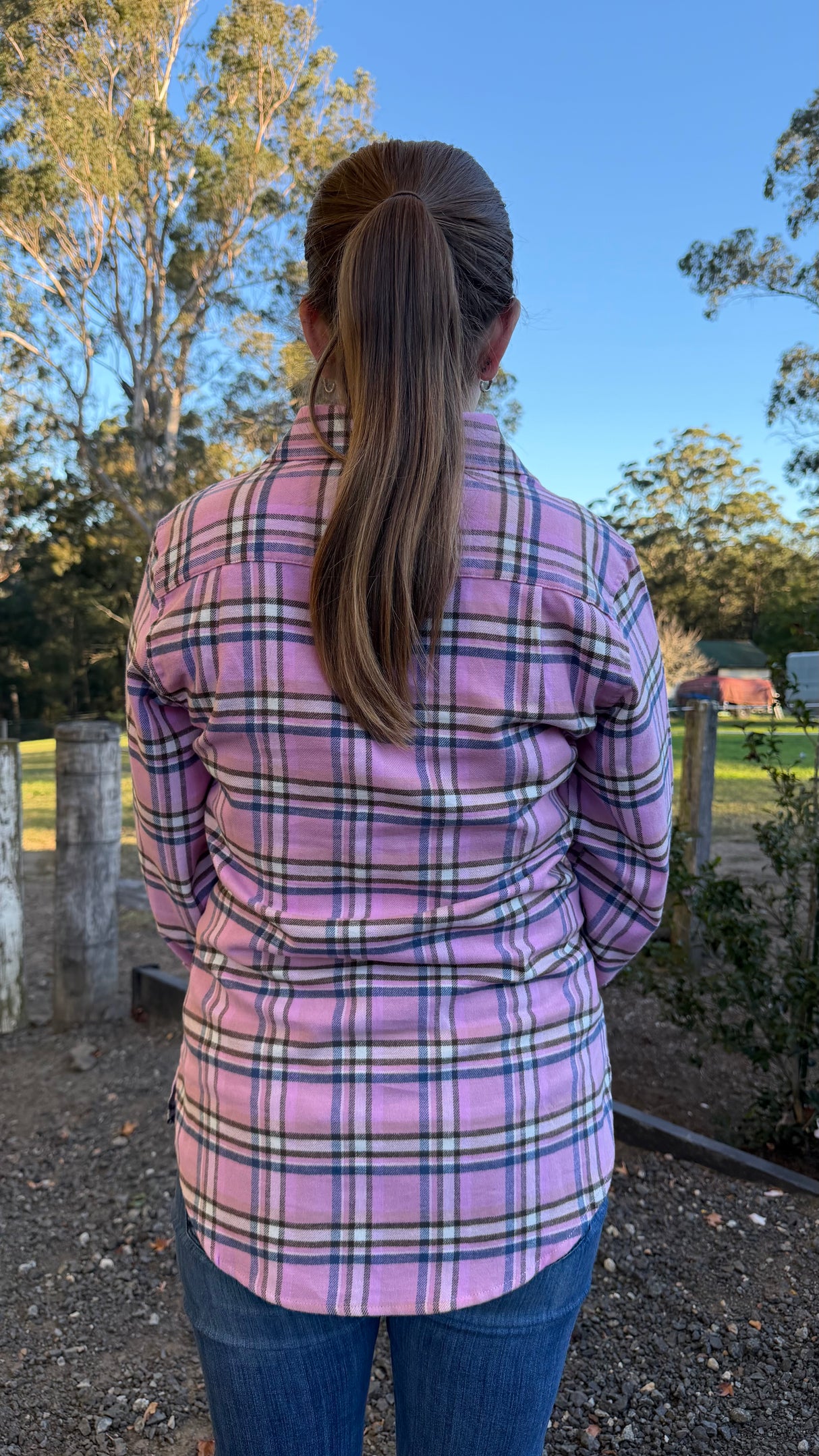 A woman viewed from behind wearing a pink plaid flannel shirt and blue jeans, standing outdoors in a rural setting with trees, a fence, and a blue sky.