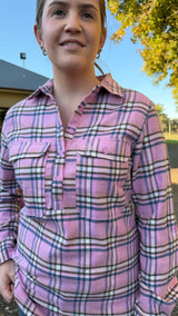 A woman wearing a pink plaid button-up shirt with purple and white stripes, photographed outdoors against a background of blue sky and green foliage.