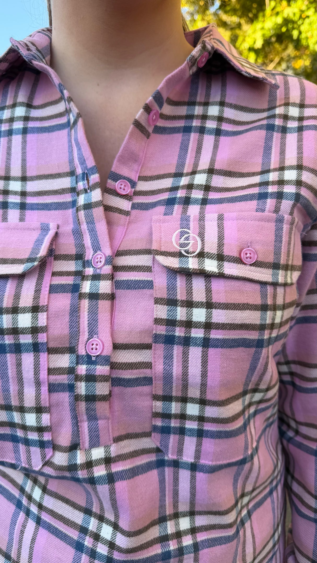 A close-up of a pink plaid button-up shirt worn by a person, featuring pink buttons and a white embroidered logo on the chest pocket.