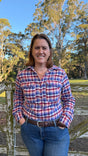 A smiling woman wearing a red, blue, and white plaid long-sleeve button-up shirt and blue jeans, standing outdoors in front of a rustic wooden fence and trees under a blue sky.