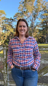 A smiling woman wearing a red, blue, and white plaid long-sleeve button-up shirt and blue jeans, standing outdoors in front of a rustic wooden fence and trees under a blue sky.