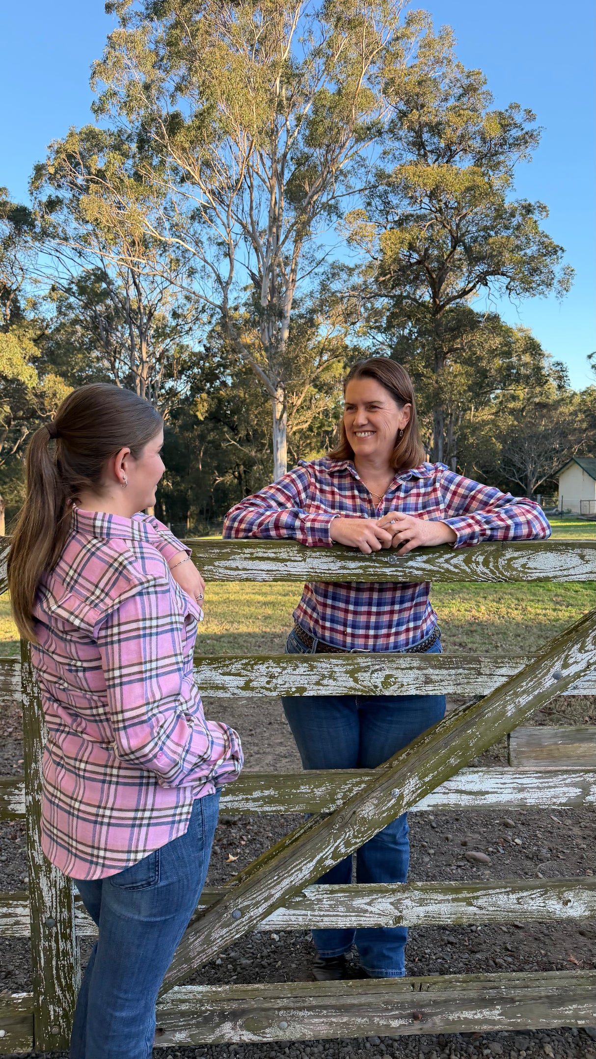 Two women in matching pink, red, and blue plaid flannel shirts and jeans standing outdoors on a rustic wooden fence with trees and blue sky in the background.