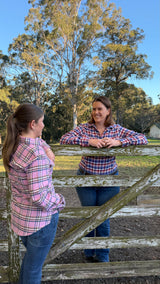 Two women in matching pink, red, and blue plaid flannel shirts and jeans standing outdoors on a rustic wooden fence with trees and blue sky in the background.