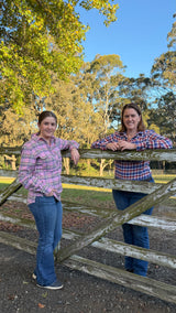 Two women standing outdoors behind a rustic wooden fence, wearing matching pink and blue plaid button-up shirts and blue jeans, with trees and blue sky in the background.